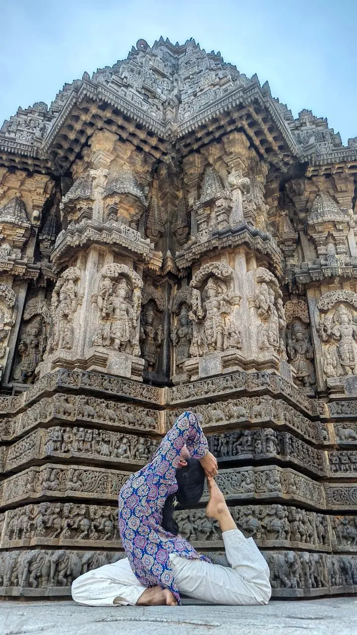 नक्काशीदार होयसल मंदिर की दीवार के सामने बैठकर किया गया गहरा पीछे झुकने वाला आसन